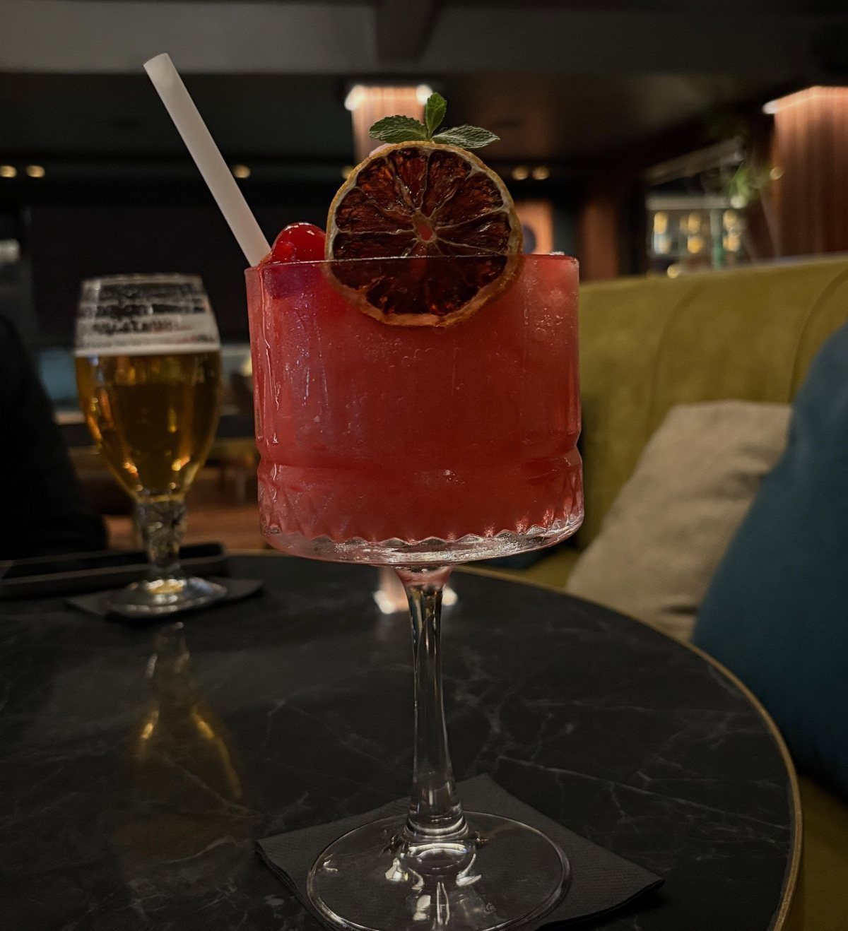 Red cocktail with orange garnish and a beer on a dark marble table.