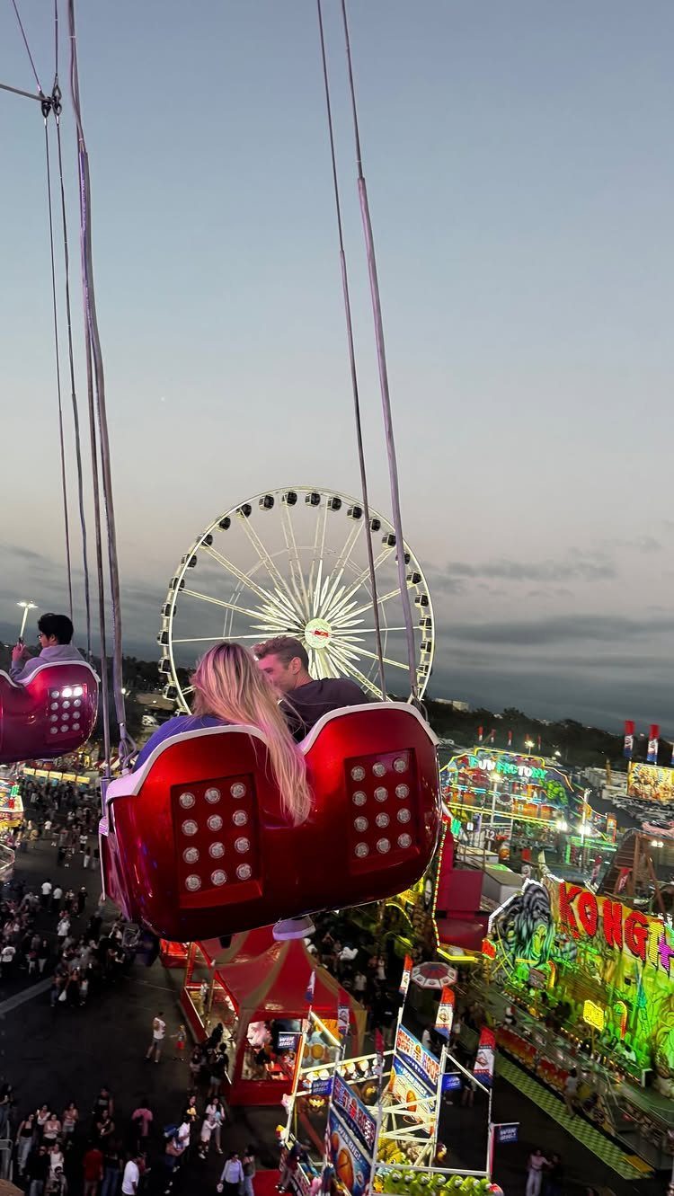 Couple in a red ride carriage at a carnival with a Ferris wheel background.