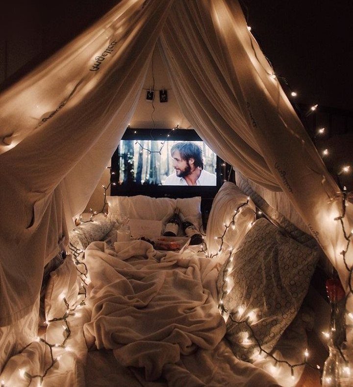 Cozy indoor blanket fort with string lights and a movie playing on screen.