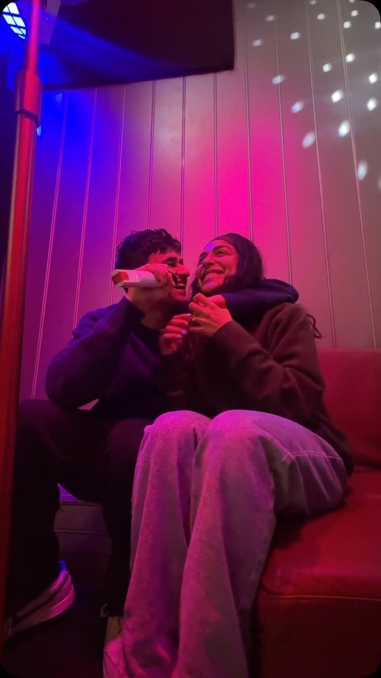 Couple laughing and singing into a microphone in a neon-lit karaoke room.