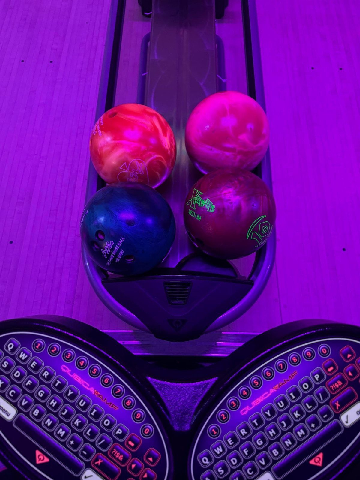 Colorful bowling balls in a return rack under purple neon bowling alley lights.