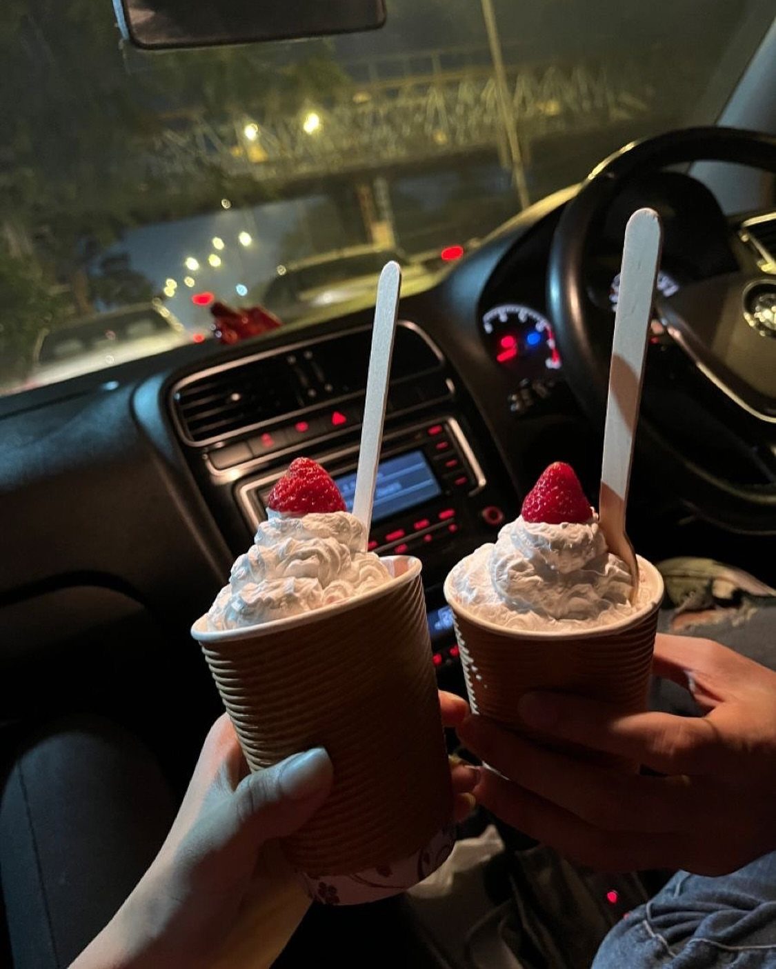 Two strawberry whipped cream drinks held inside a car during a night drive.
