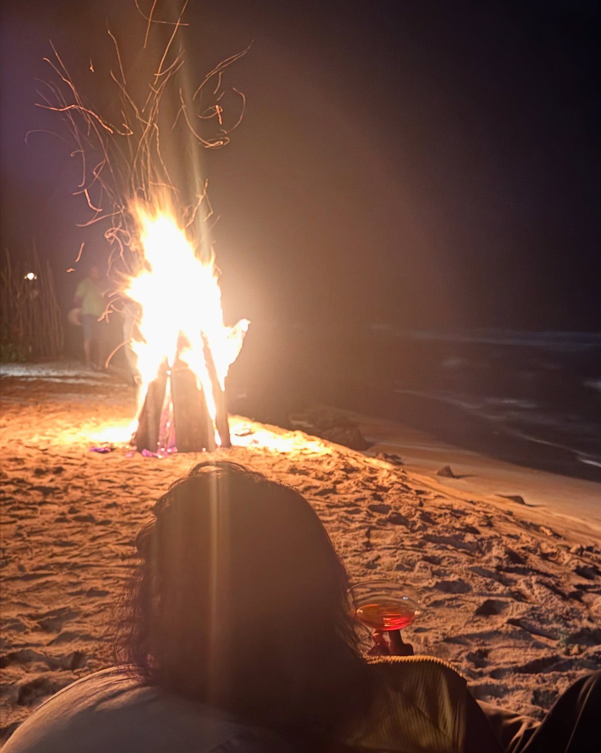 Person sitting on a sandy beach at night watching a large glowing bonfire.