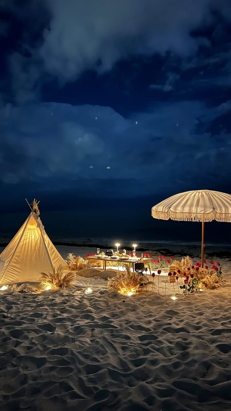Romantic beach dinner setup with a low table, teepee, and umbrella at night.