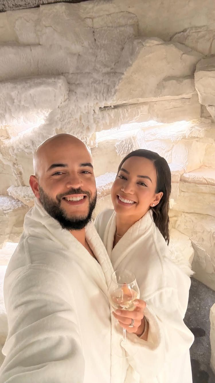 Smiling couple in white bathrobes posing in a white textured salt cave room.