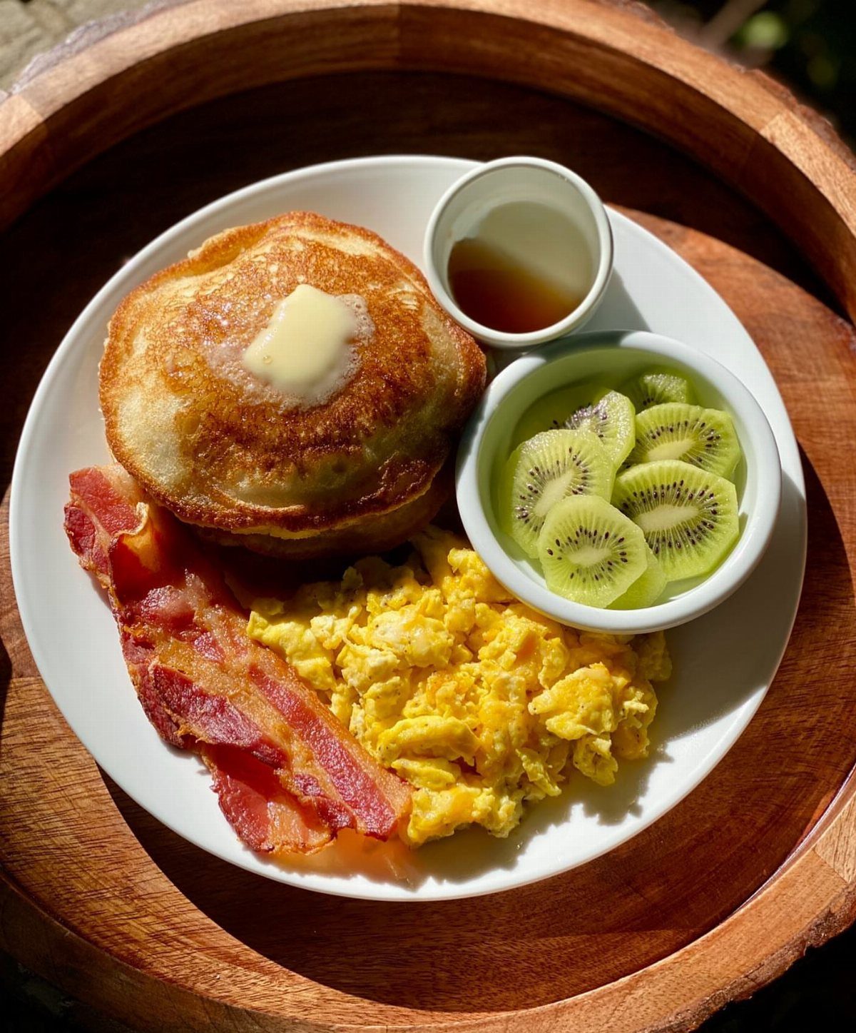 Breakfast plate with pancakes, scrambled eggs, bacon, syrup, and sliced kiwi on wood.
