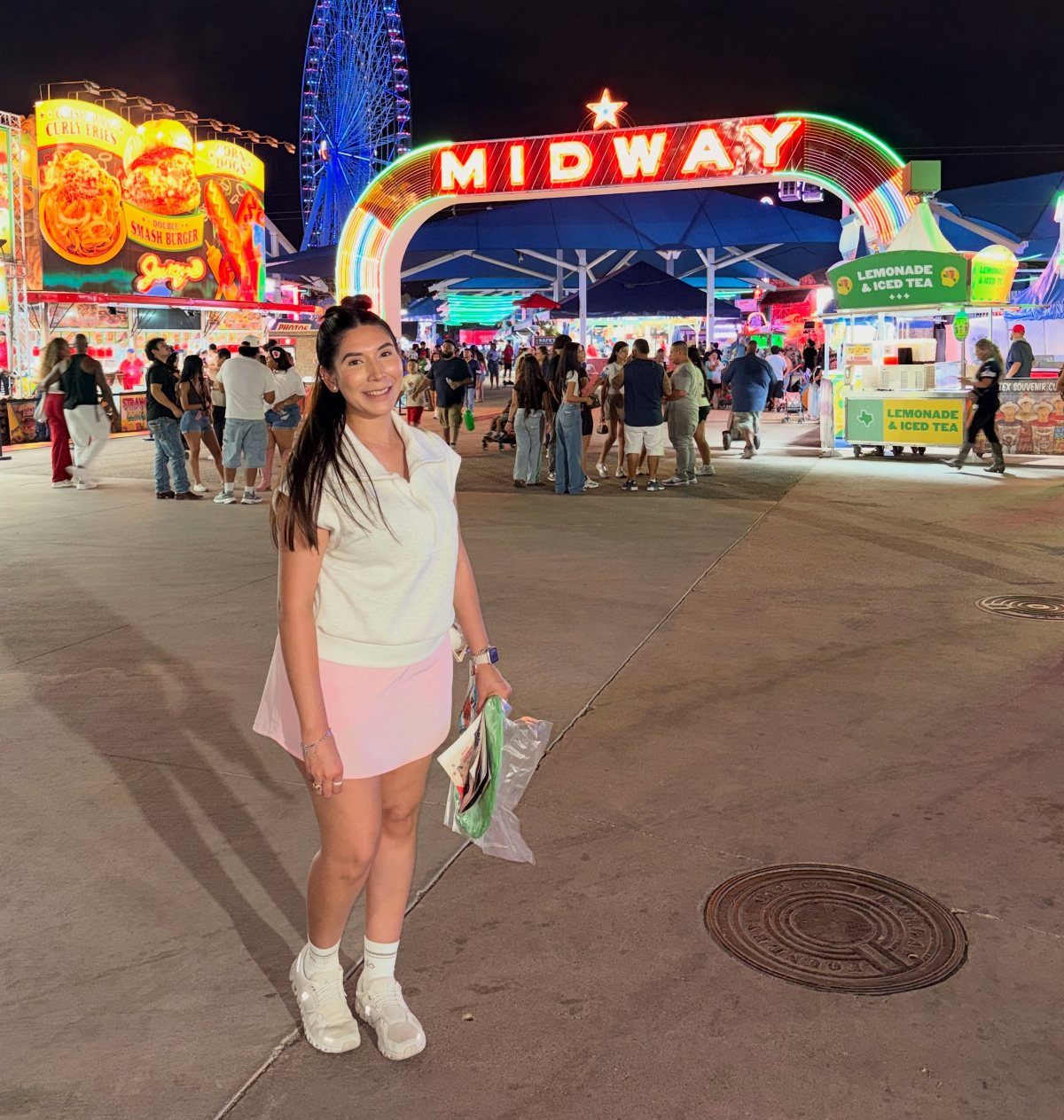 Woman standing in a carnival midway with a neon sign and Ferris wheel behind.