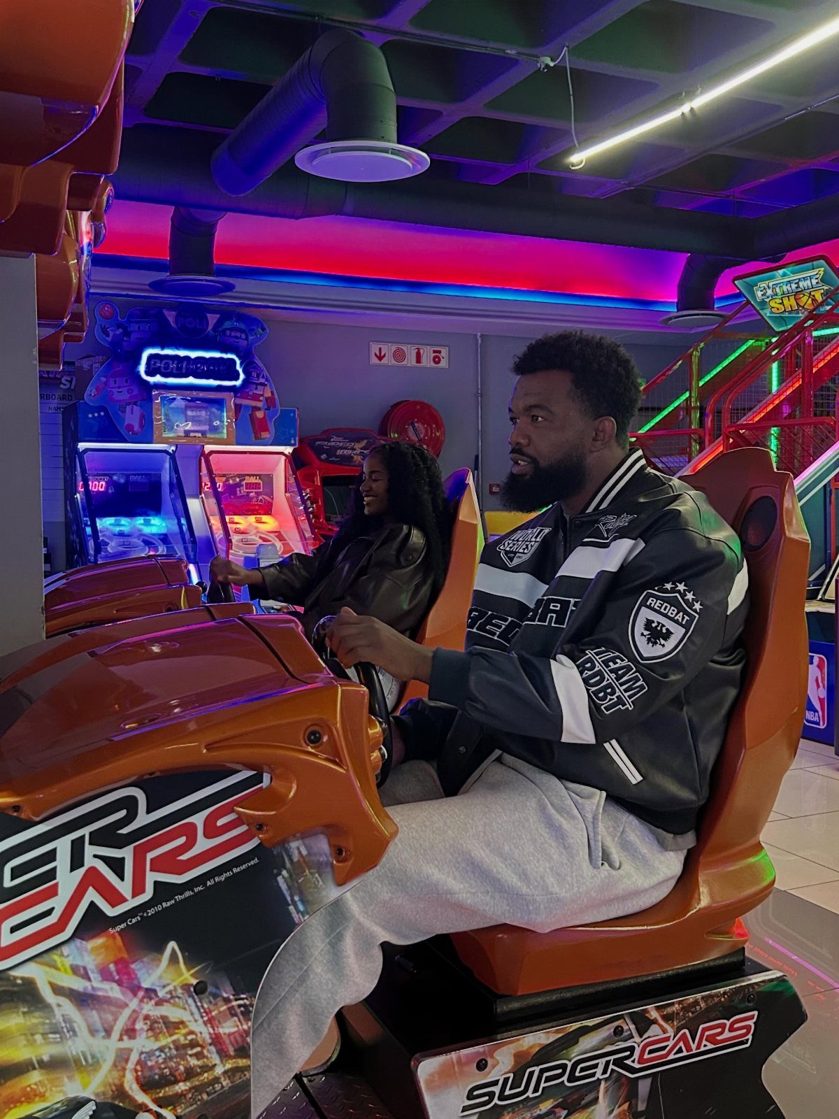 A couple playing racing simulator games in a neon-lit arcade center.