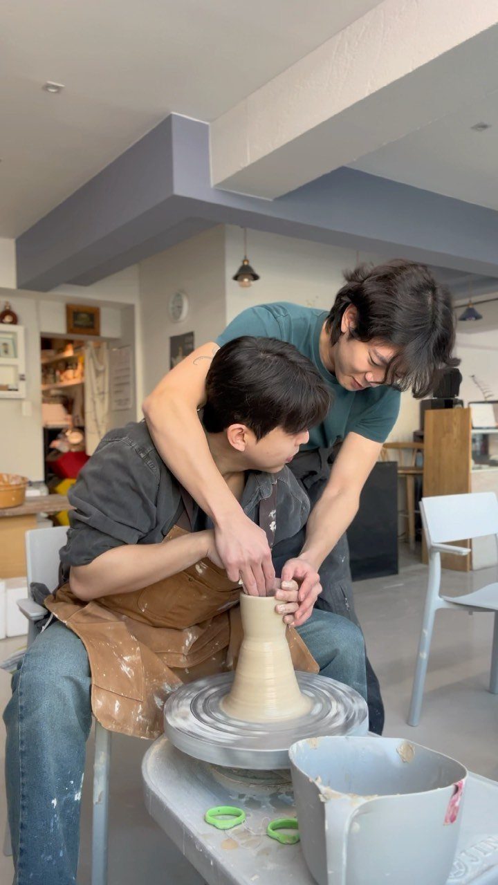 Two men working together to shape a clay vase on a pottery wheel.