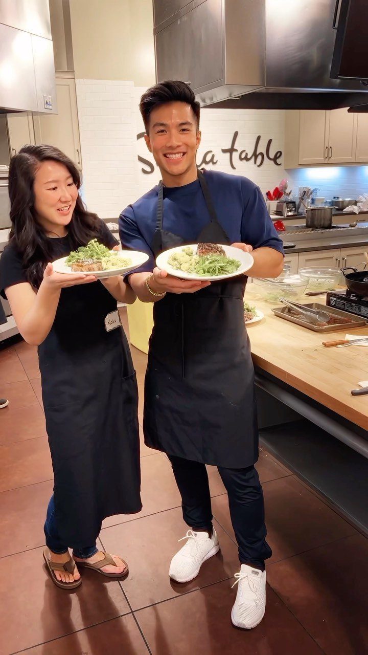 Couple in black aprons holding plates of food in a professional kitchen
