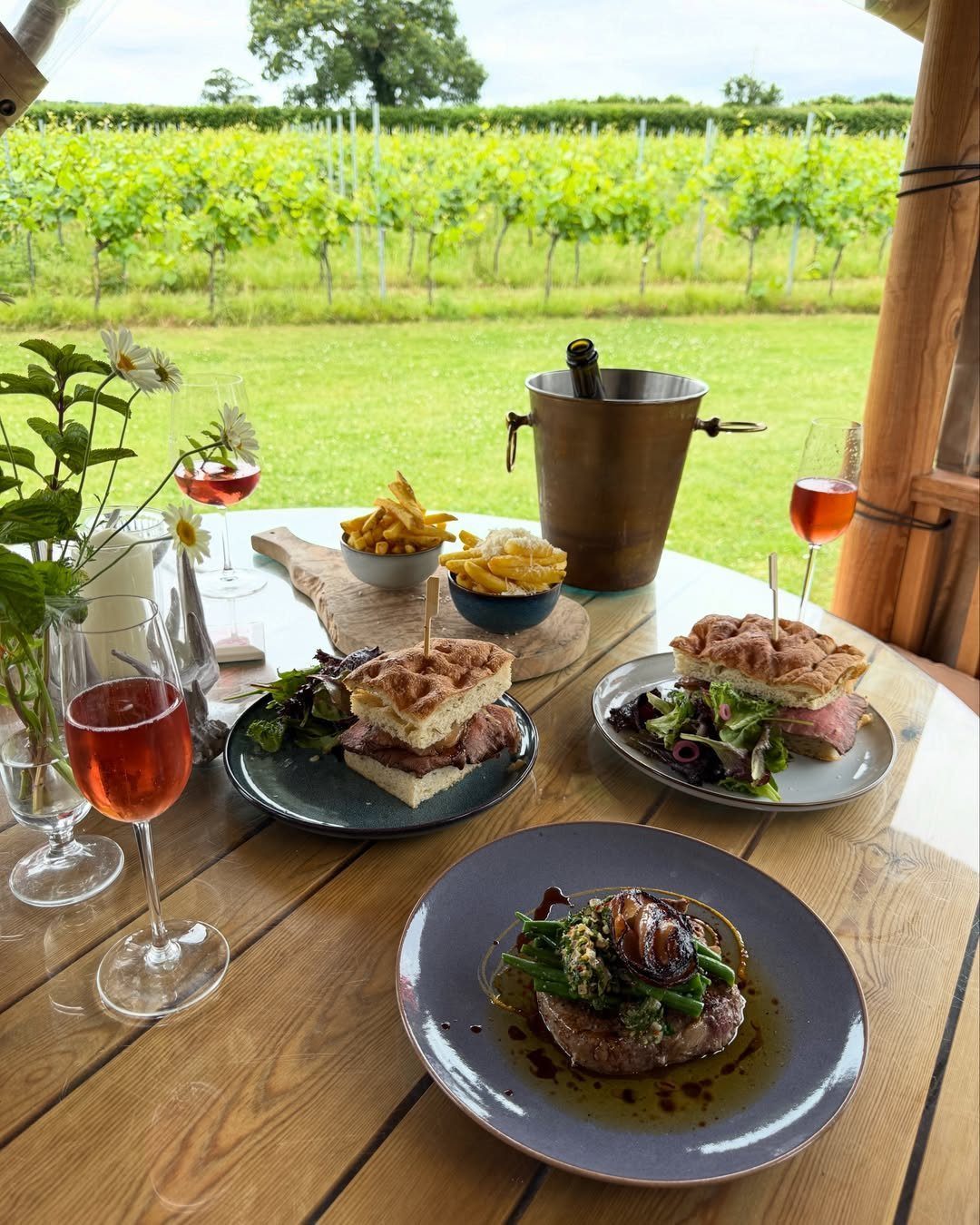Outdoor table set with wine, sandwiches, and steak overlooking a green vineyard.