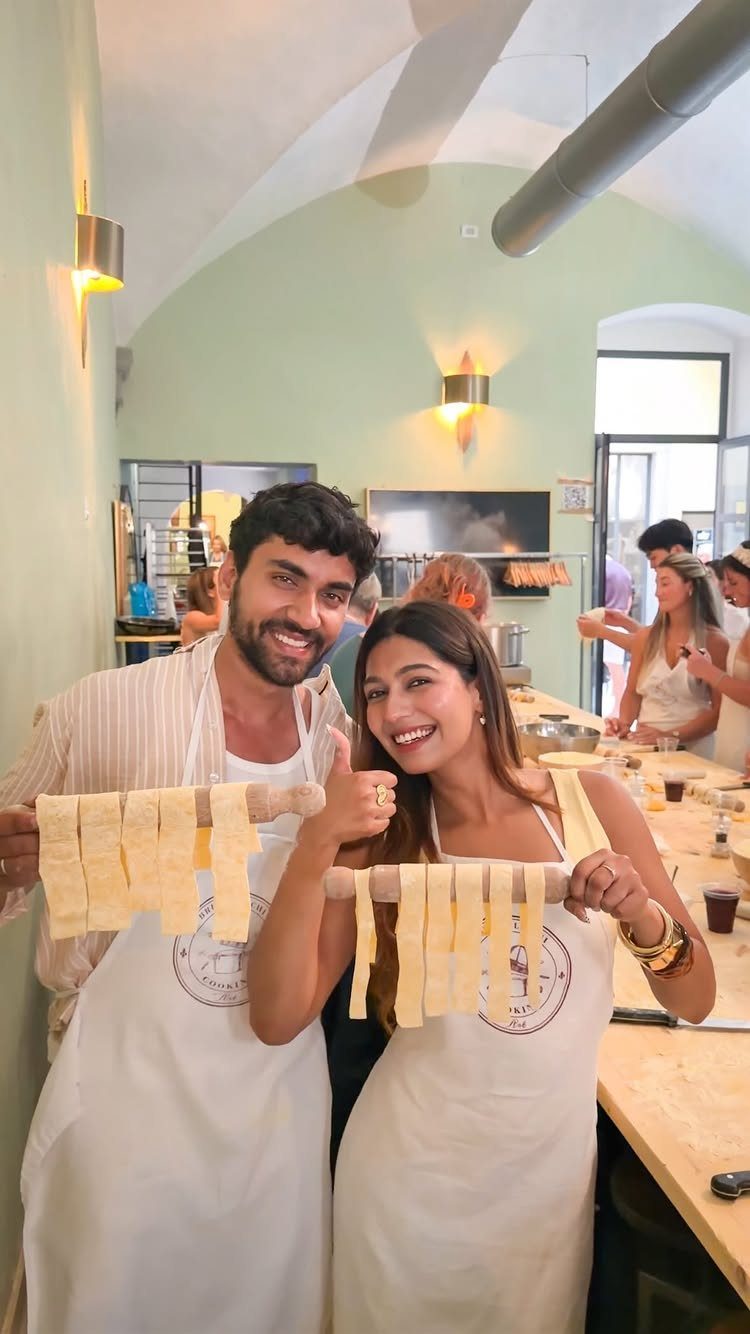 Smiling couple in white aprons holding rolling pins with fresh pasta strips.