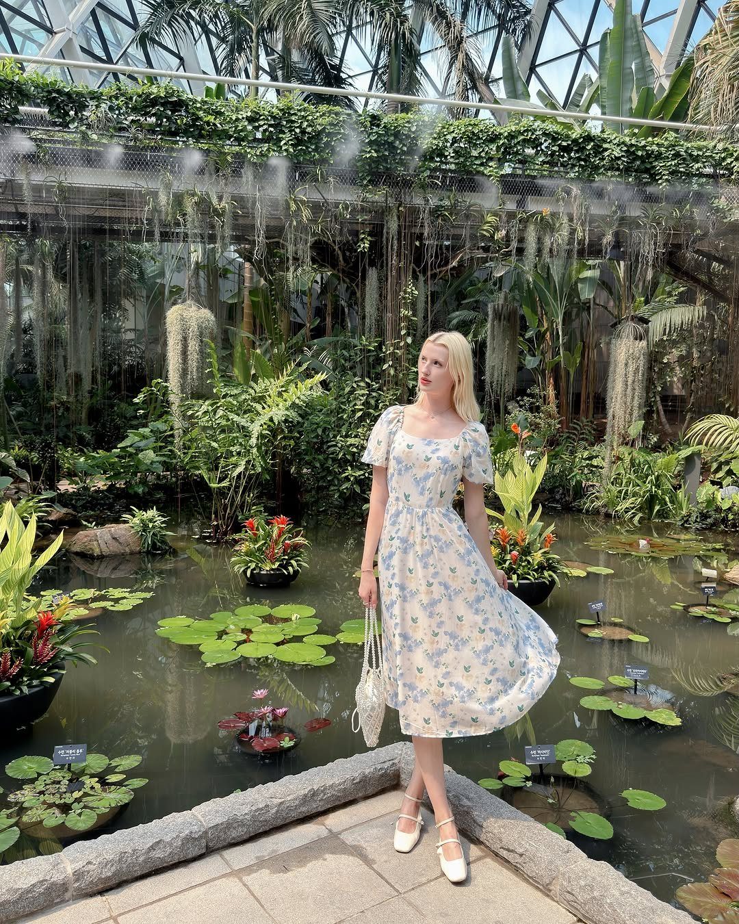 Woman in floral dress standing in a lush botanical garden conservatory by a pond.