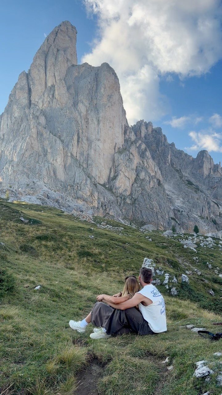 Couple sitting on a grassy slope looking at a towering mountain peak.