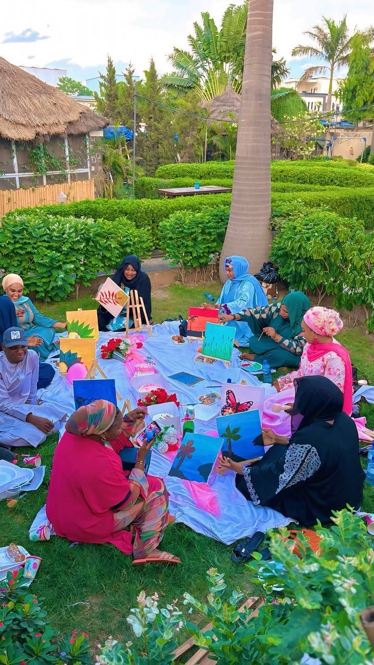 People sitting on a lawn painting colorful landscapes on canvases during an outdoor session.