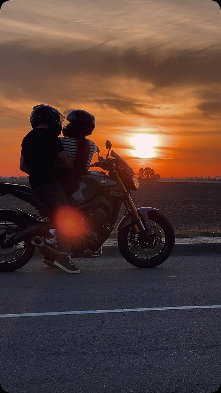 Couple on a motorcycle silhouetted against a vibrant orange sunset over a horizon.