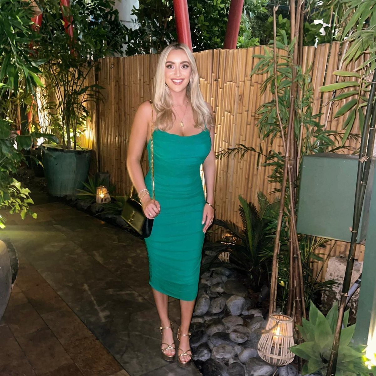 Woman in a tight emerald green midi dress and gold heels standing outdoors.