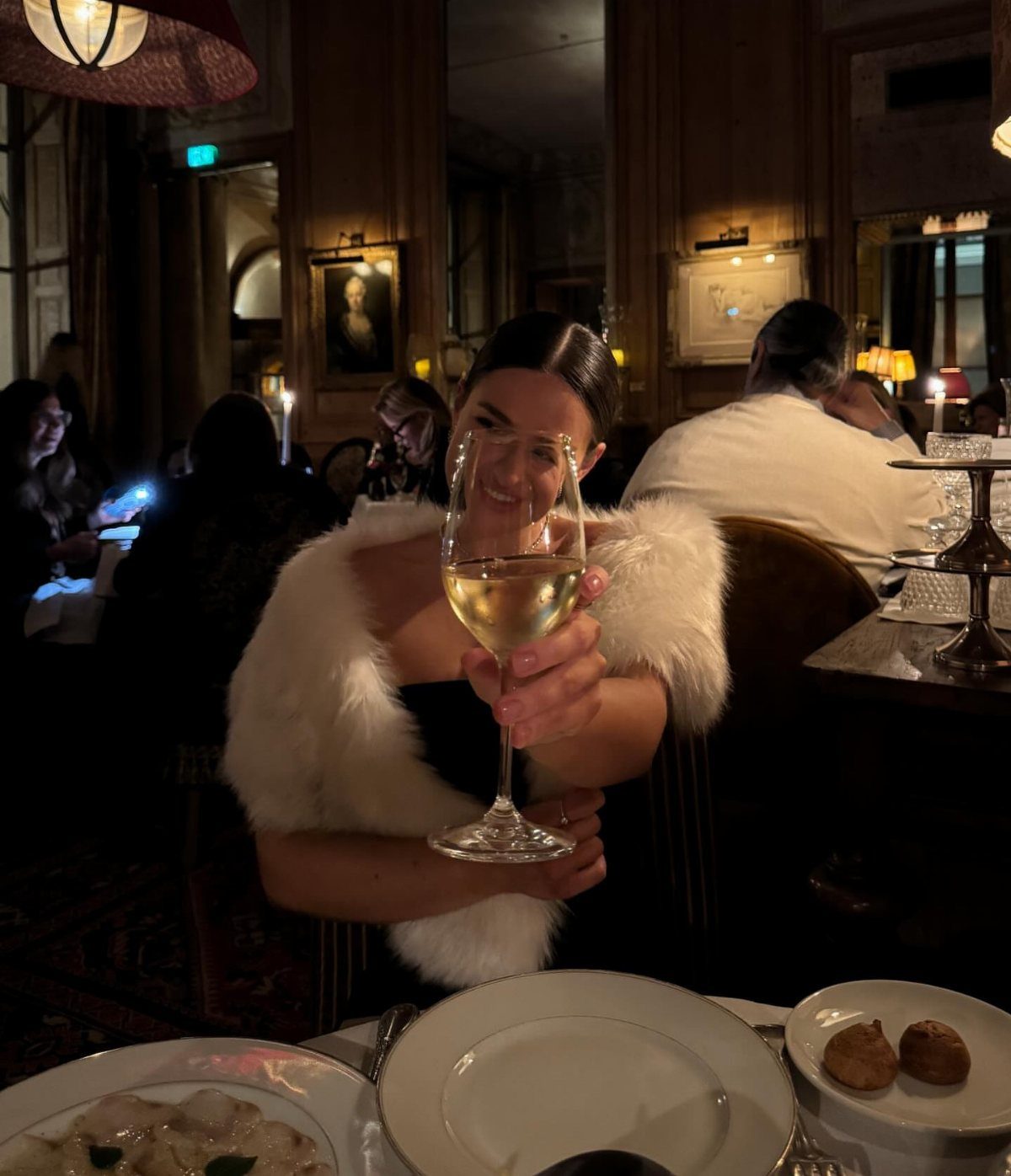 Woman in a dark dress and white fur stole holding a wine glass.