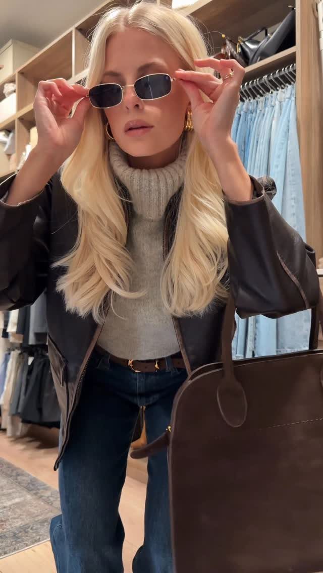 Blonde woman in a leather jacket, grey turtleneck, and jeans in a store.