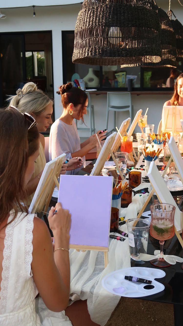 Women painting on small canvases at an outdoor table with art supplies and drinks.
