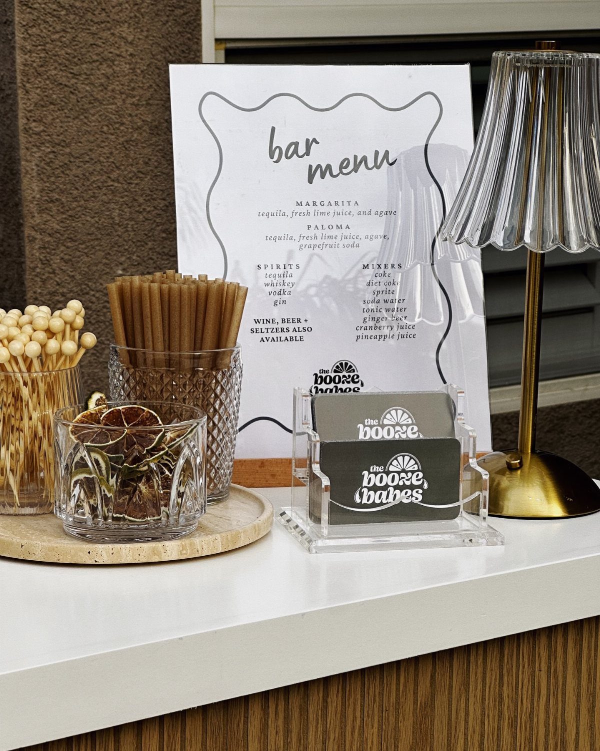 Bar menu and cocktail accessories on a white counter with a gold lamp.