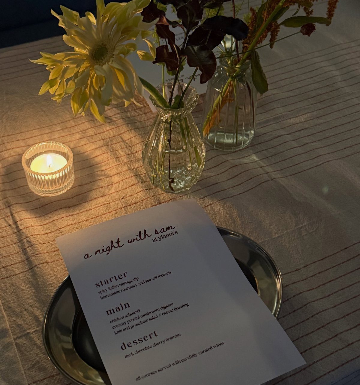 Dinner table setting with a printed menu, lit candle, and floral arrangements.