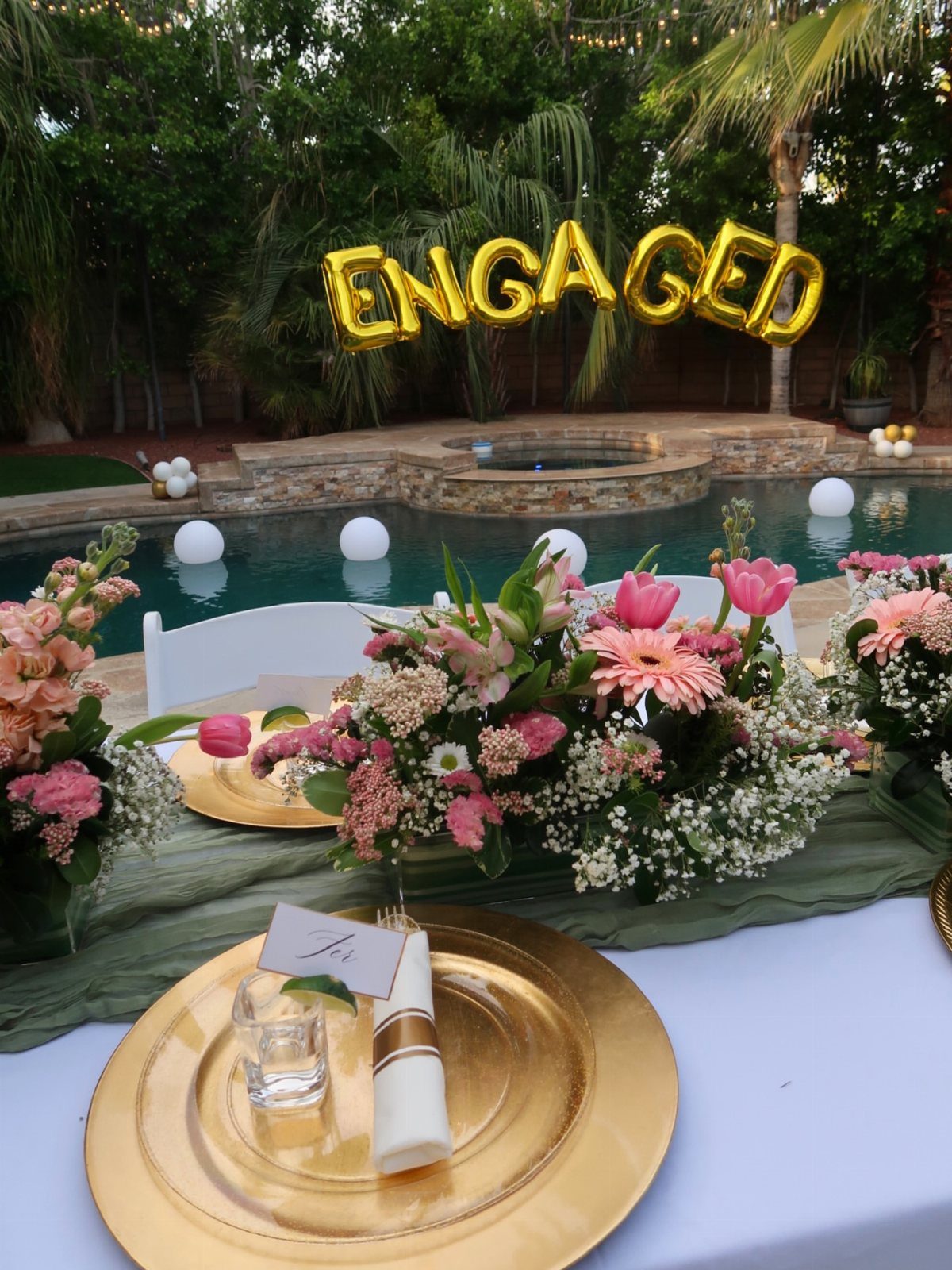 Poolside engagement party table with gold plates, pink flowers, and ENGAGED balloons.