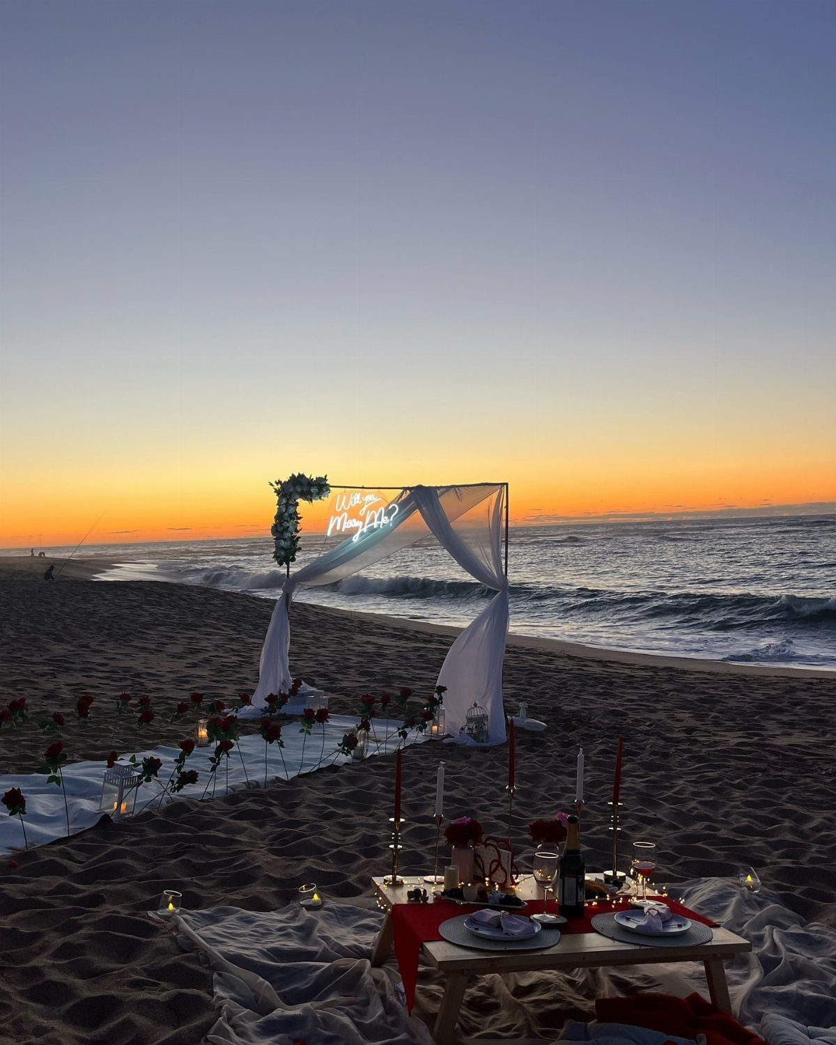 Beach picnic setup with low table, red roses, and white arch at sunset.