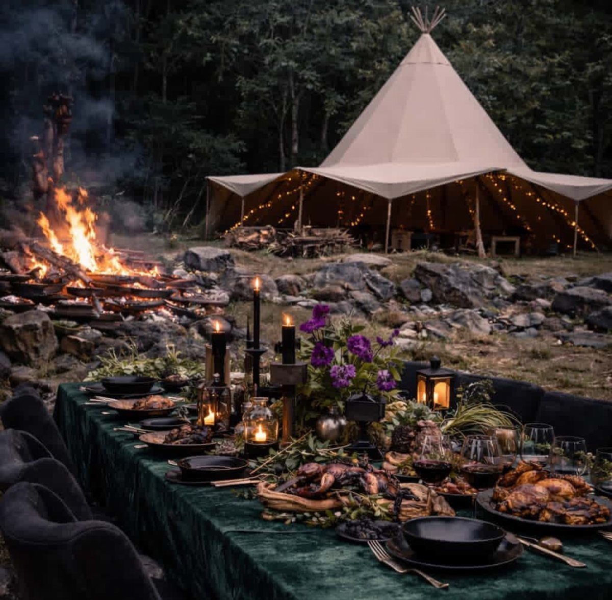 Outdoor dinner table with roasted food near a bonfire and lit teepee tent