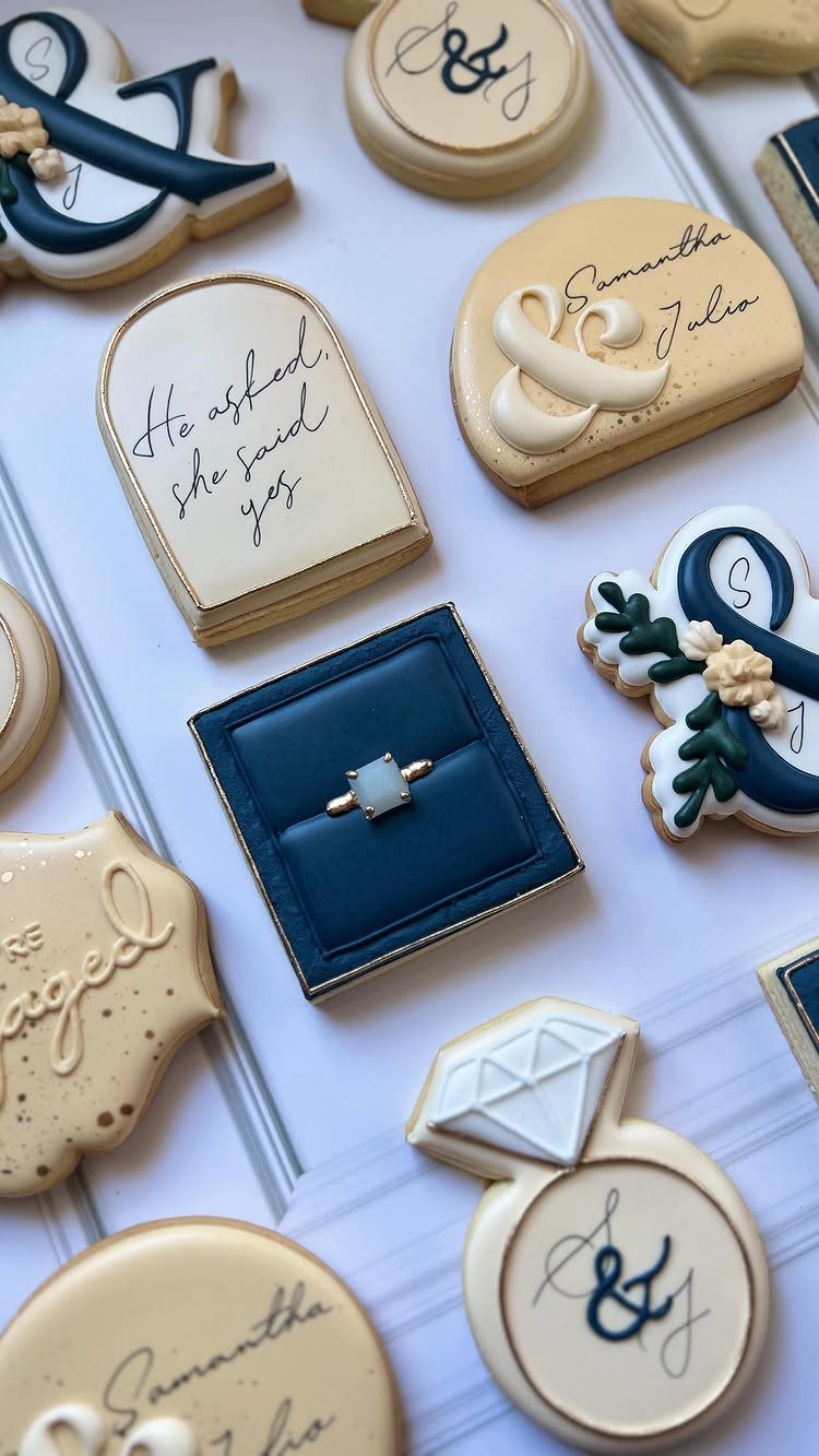 Decorated engagement cookies featuring a diamond ring, ring box, and elegant cursive lettering.