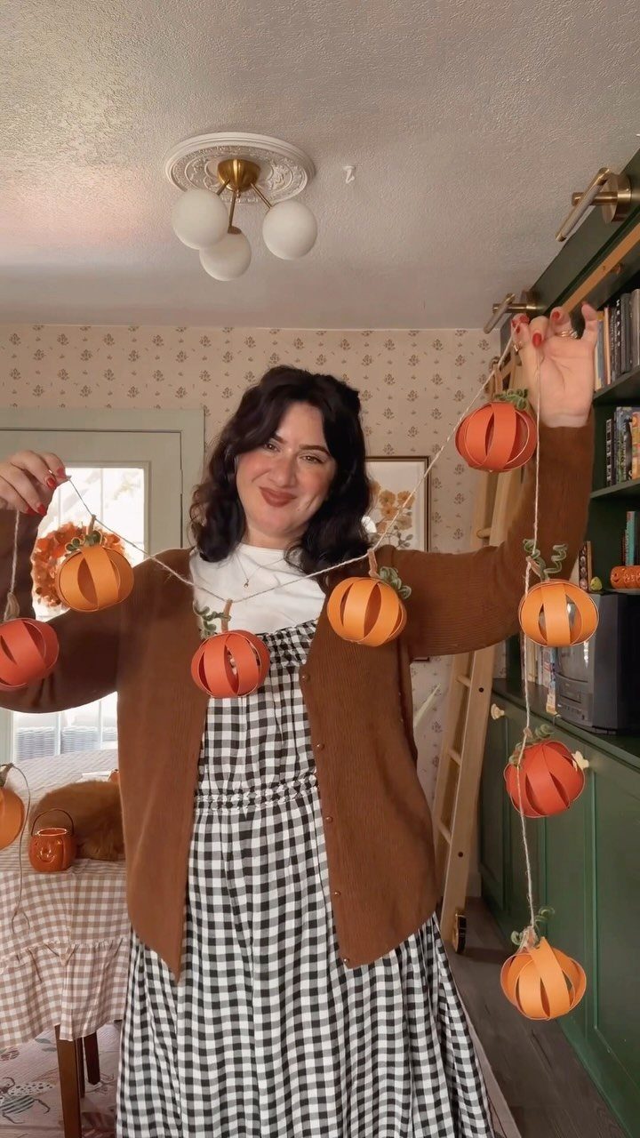 Woman in autumn attire holding a string of orange pumpkin decorations indoors.