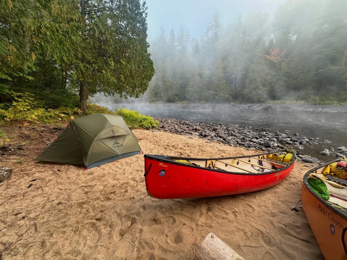 Green camping tent and red canoe on a sandy shore by a misty river