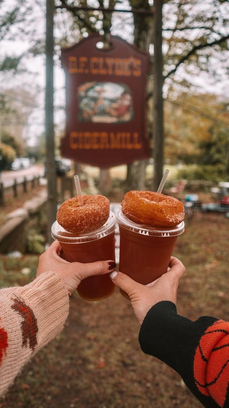 Two cider drinks topped with donuts held in front of a cidermill sign.