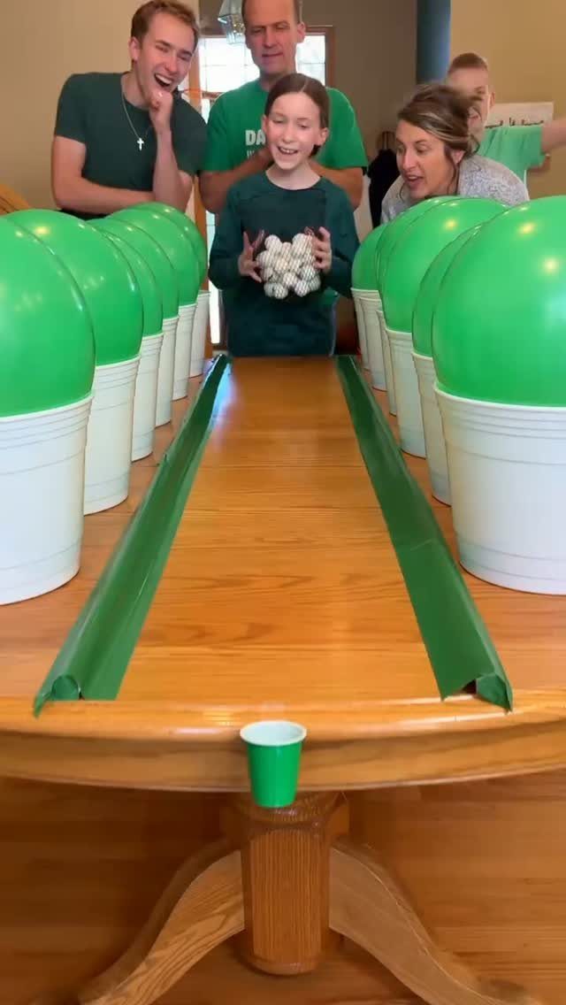 Family playing a homemade table game with green balloons and white cups.