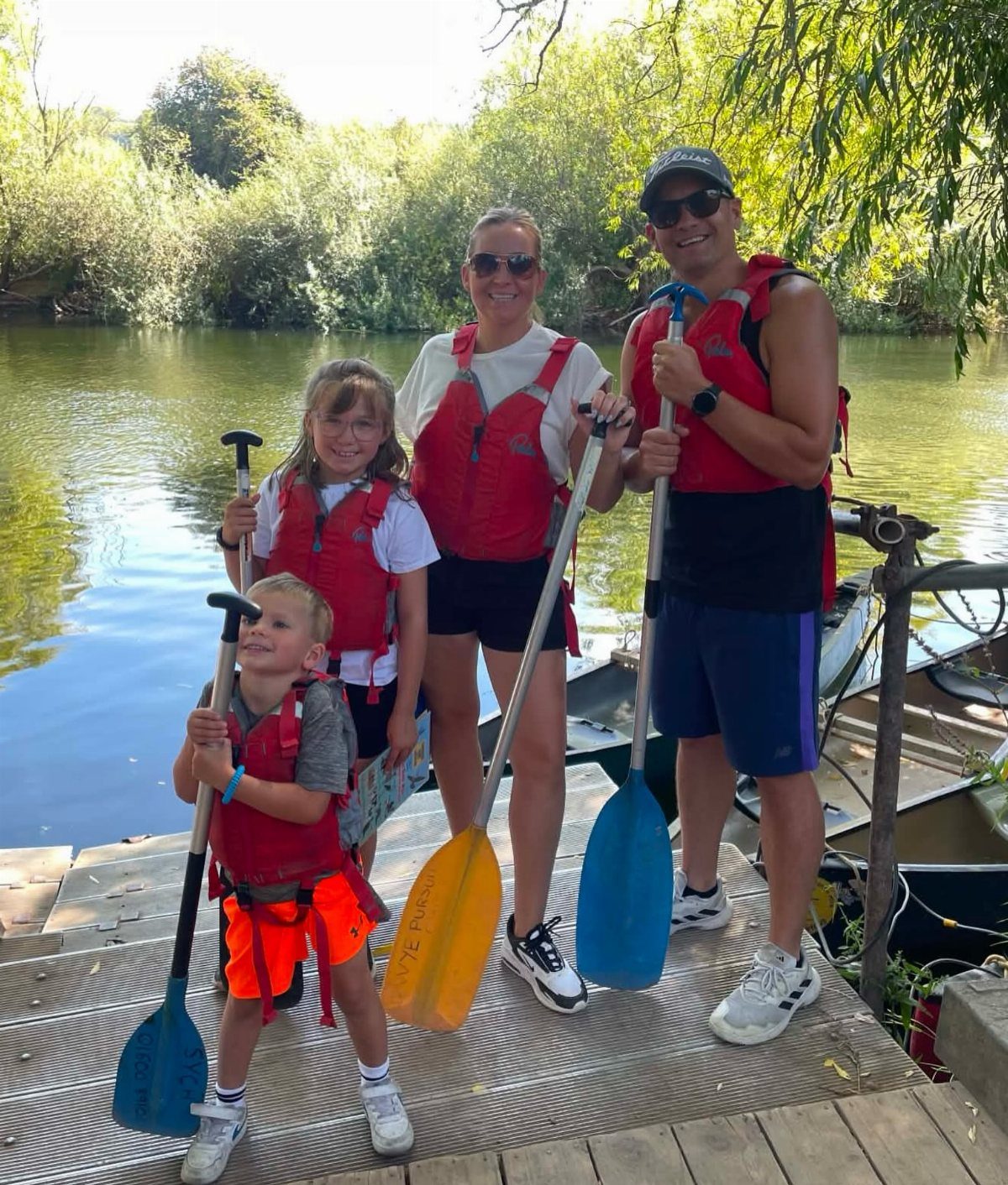 Family of four in red life jackets holding paddles on a river dock.