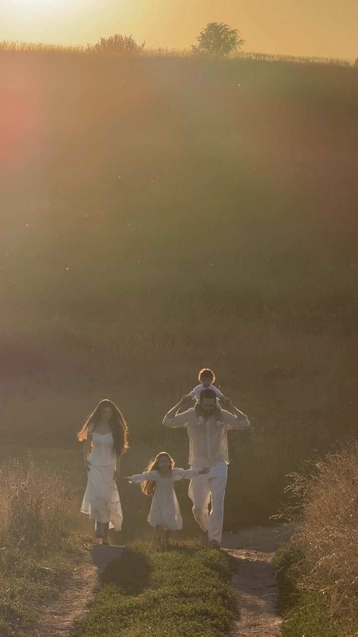 Family in white clothing walking on a dirt path through a grassy field at sunset