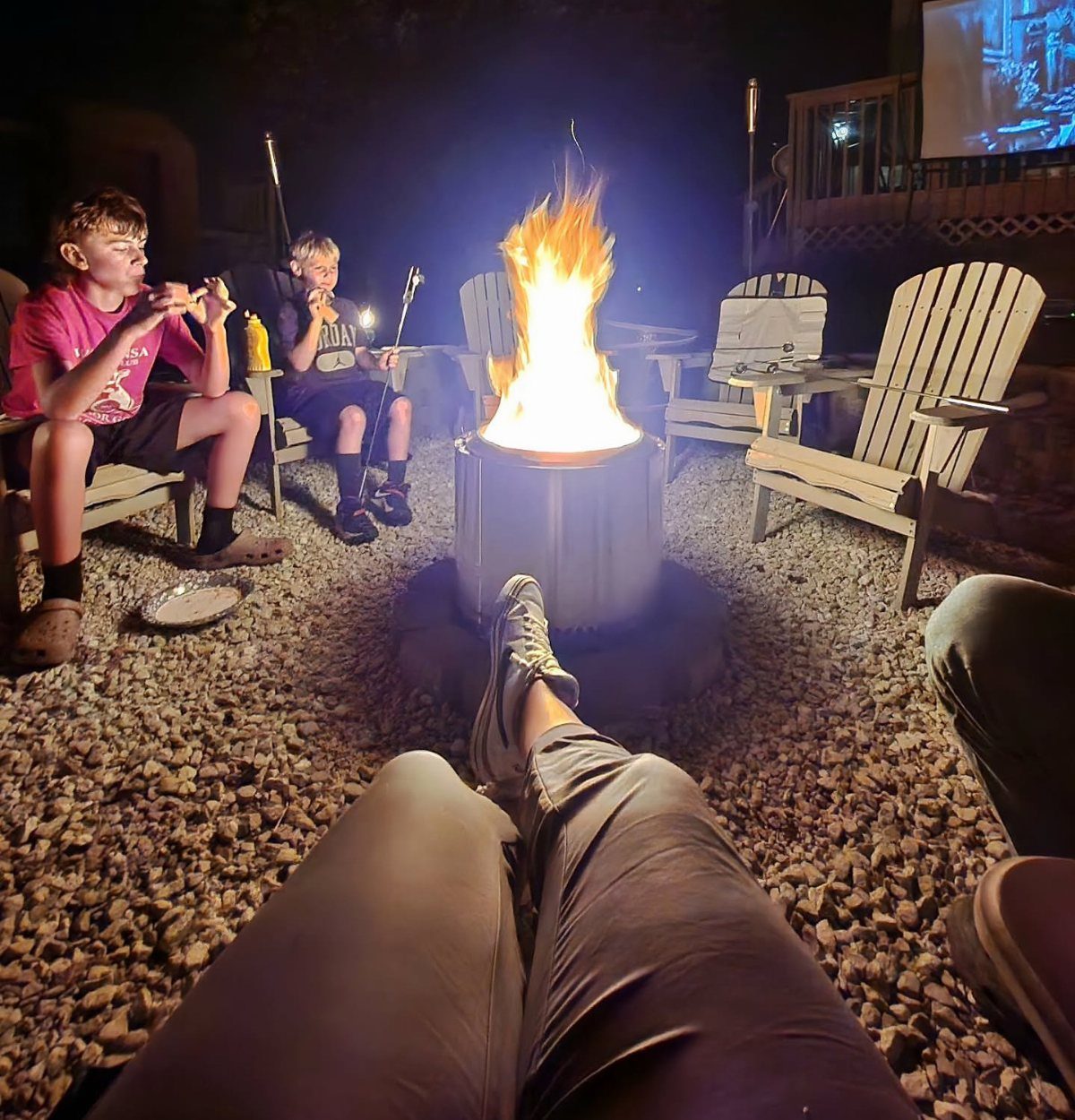 People roasting marshmallows around a bright bonfire in a metal pit at night.