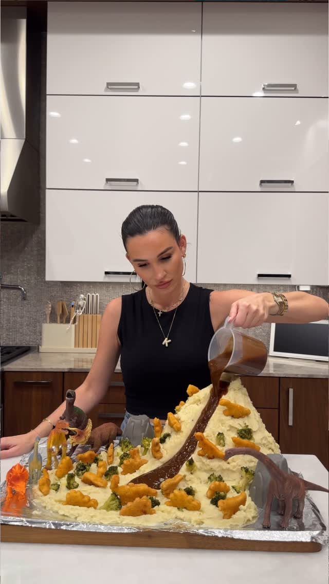 Woman pouring brown sauce over a dinosaur-themed mashed potato food landscape in kitchen