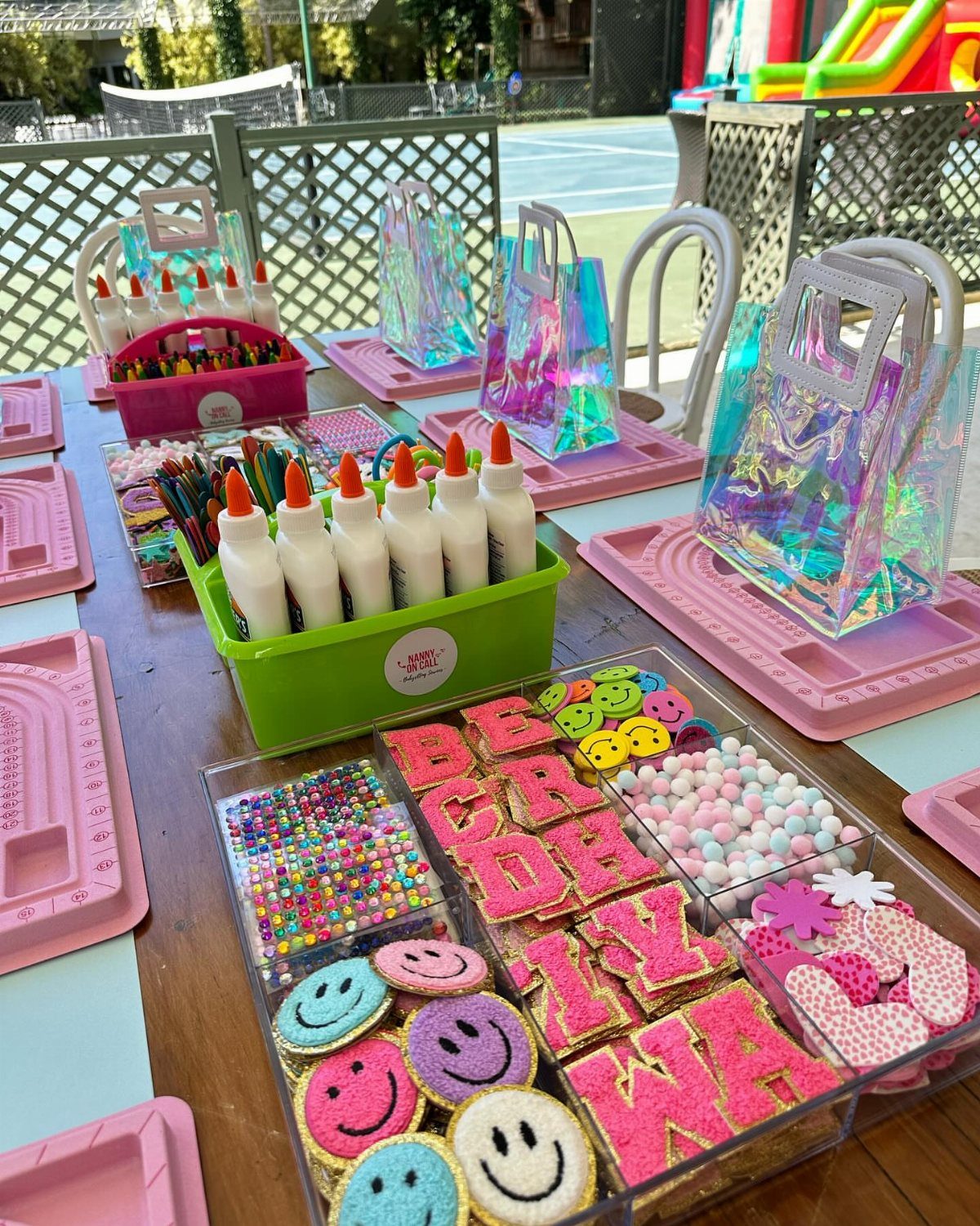 Craft station with iridescent bags, glue, markers, and glittery decorations on pink trays.