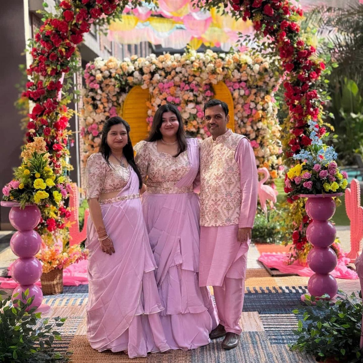 Three people in purple traditional Indian attire posing under a floral arch.