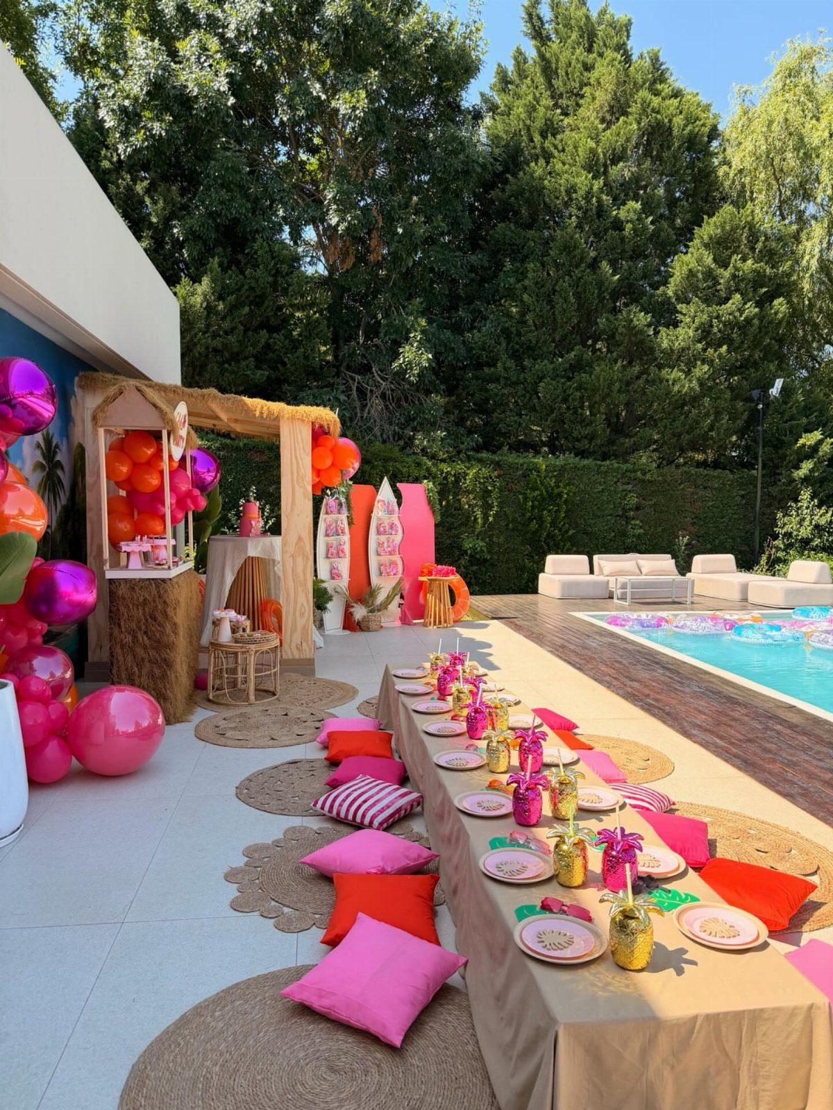 Poolside party setup with pink and orange decorations, a long table, and thatched roof.