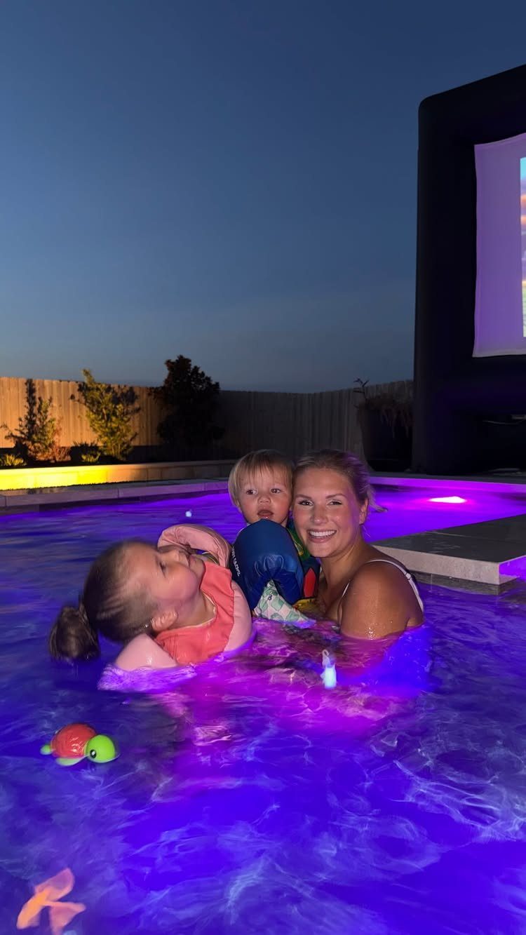 Woman and two children smiling in a purple pool with outdoor movie screen