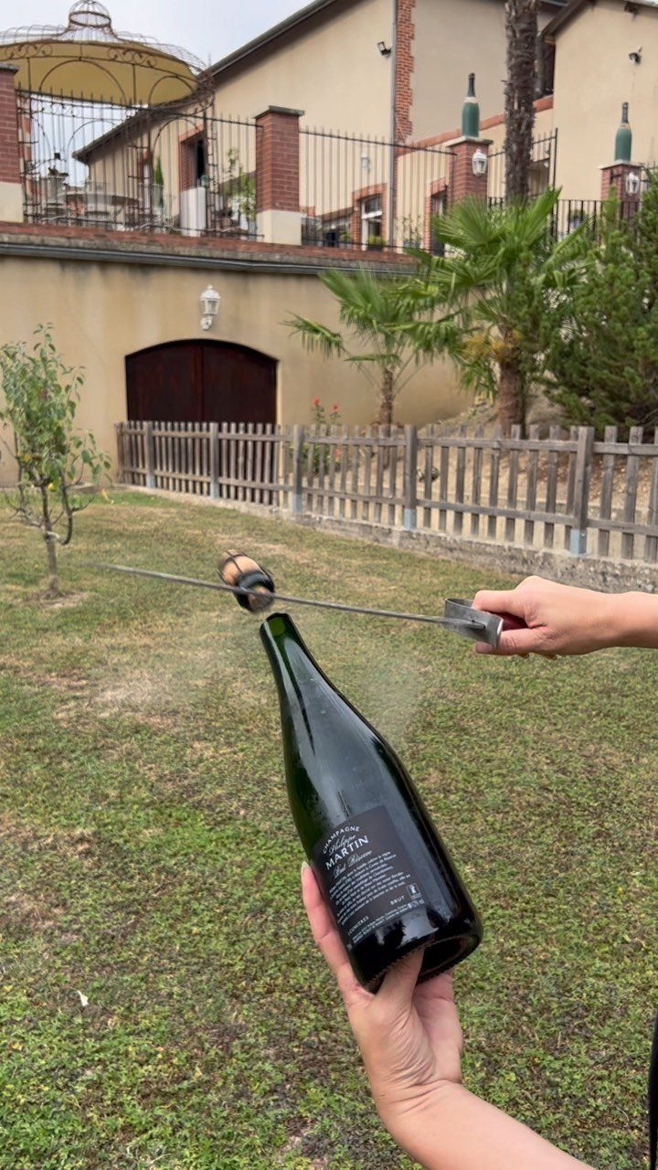 Hands sabering a champagne bottle in a grassy yard near a house