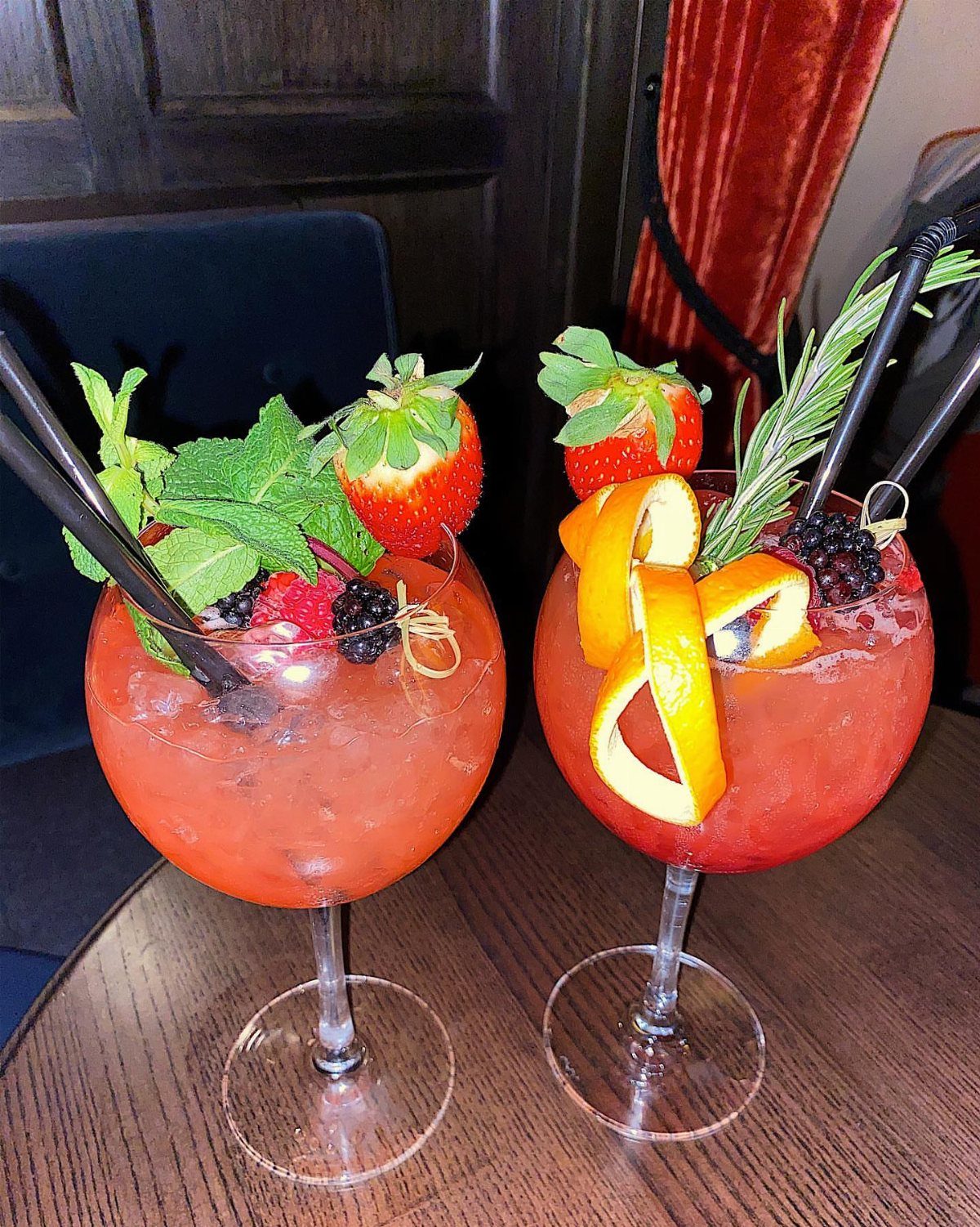 Two red cocktails with fruit garnishes on a dark wooden table in a lounge.