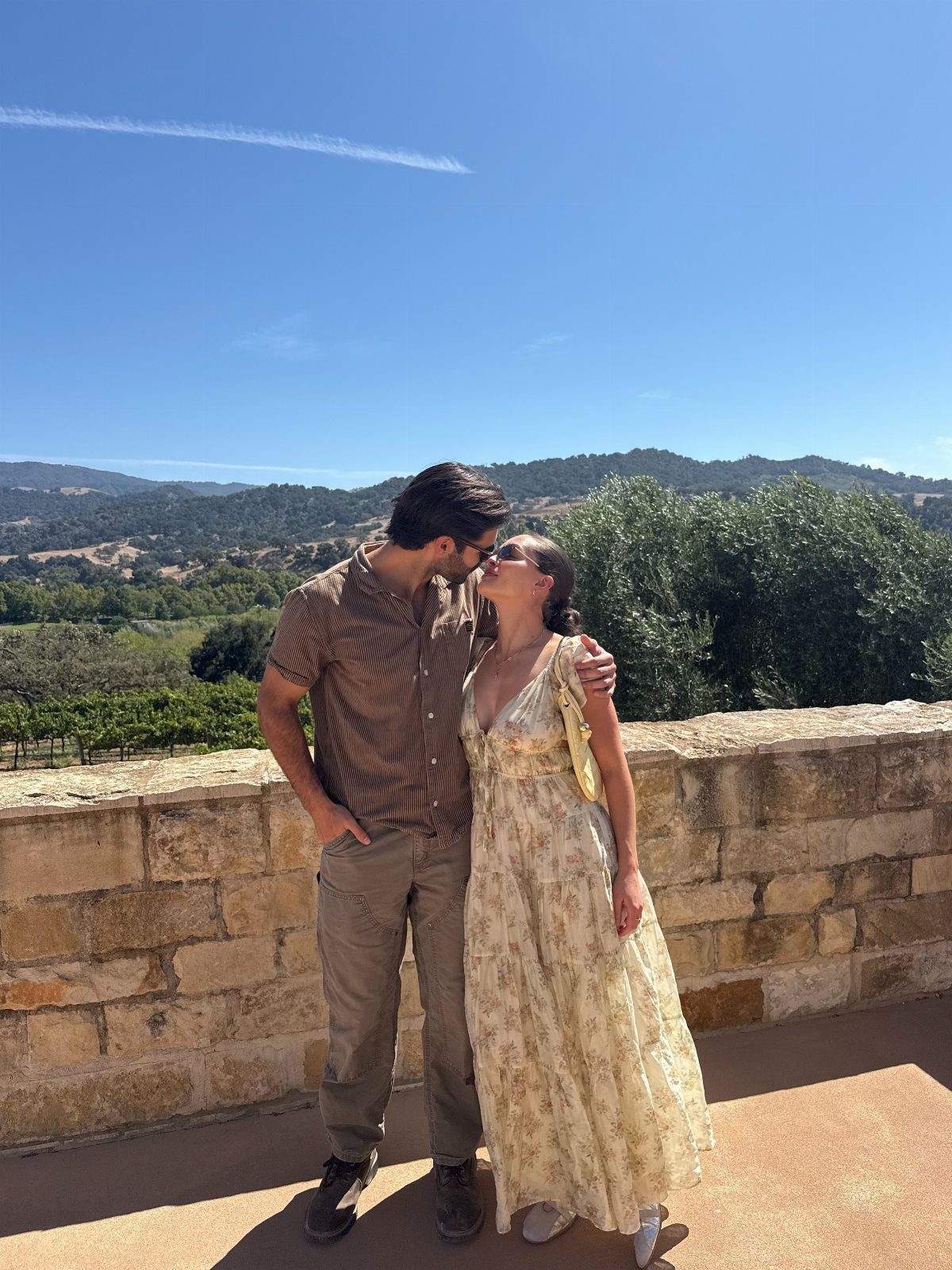 Couple in summer attire standing by a stone wall overlooking a scenic valley.