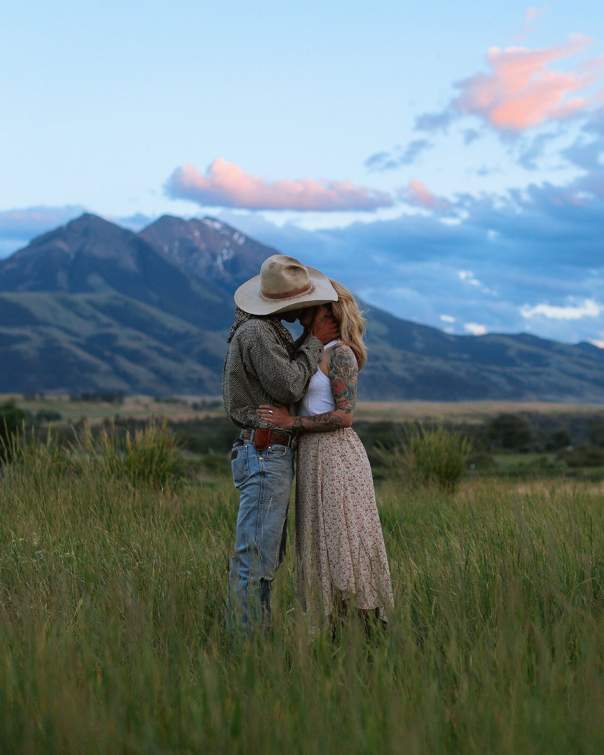 Couple kissing in a grassy field with mountains under a sunset sky.