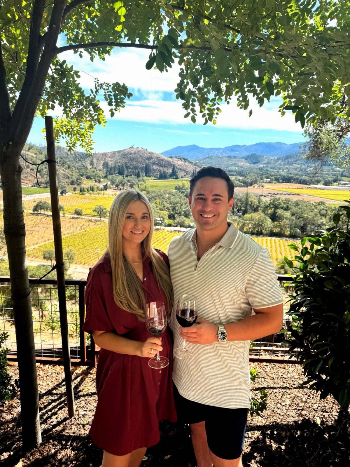 Couple holding glasses of red wine overlooking a scenic vineyard and mountains.