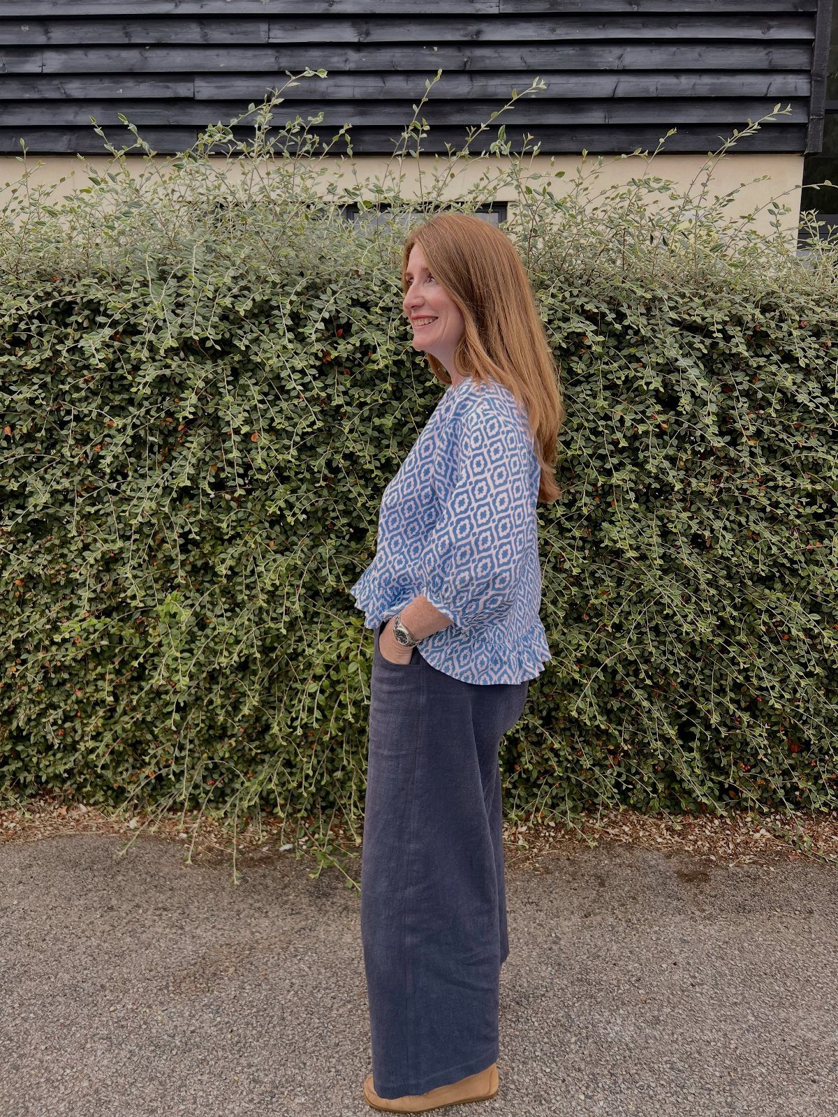 Woman wearing a blue patterned blouse and wide-leg grey trousers standing by a hedge.