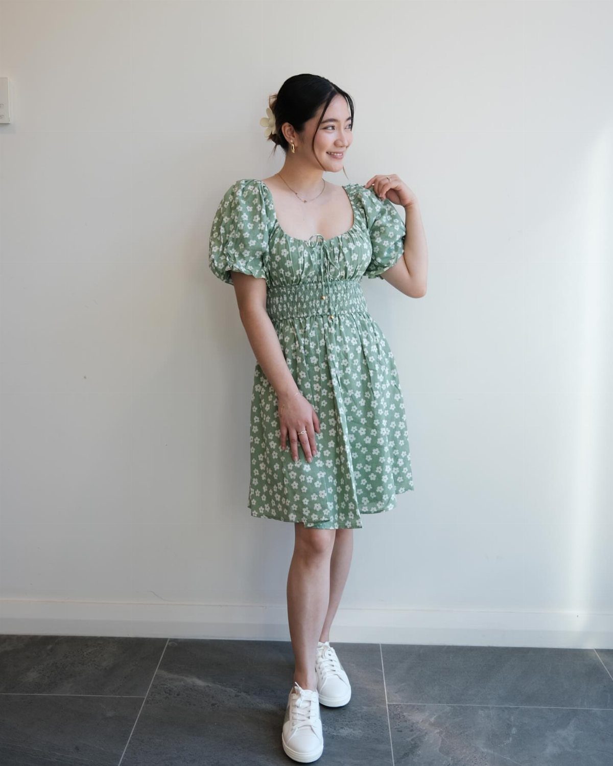 Woman wearing a green floral dress and white sneakers posing against a white wall