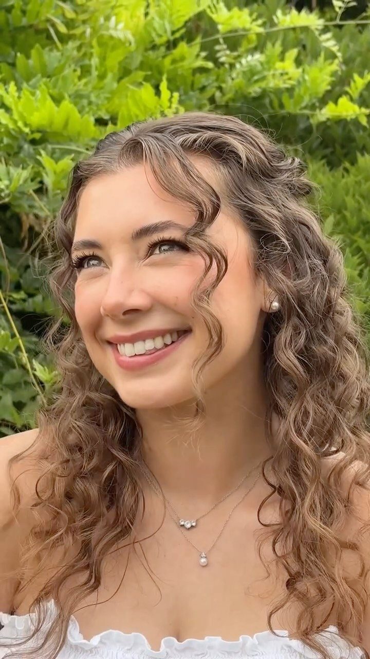 Smiling woman with curly brown hair wearing natural makeup and layered pearl necklaces.